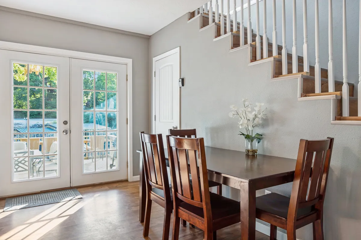 A bright dining area with a table set near French doors and a staircase, surrounded by inviting decor and natural light. The openness to the outdoors and adjacency to the living room is visible.