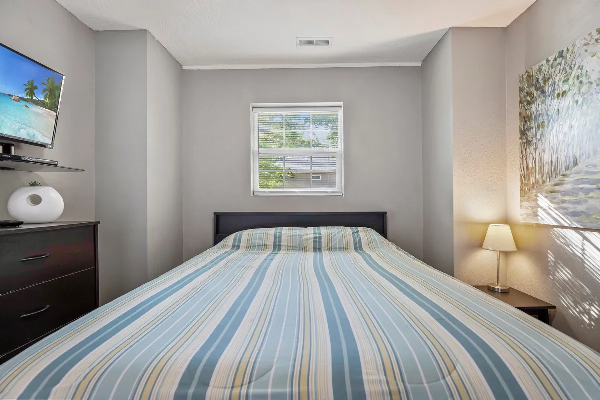 A cozy bedroom with a plush bed, TV, window letting in natural light, a bedside lamp, and artwork on the wall. The layout exudes comfort and privacy, offering a serene place to relax or enjoy a quiet movie night.