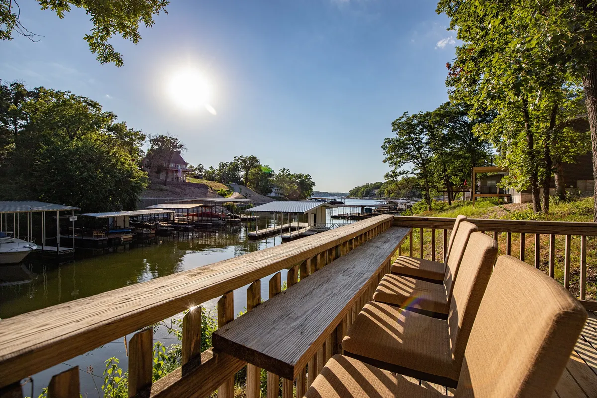 The image showcases a serene waterfront setting with a private dock stretching out over calm lake waters, boats moored nearby, and sunlight glimmering on the surface. The tranquil scene suggests a perfect spot for boating, fishing, or simply unwinding while taking in sunrise views. A spacious deck overlooks the water, hinting at opportunities for barbecues and lazy afternoons by the lake.