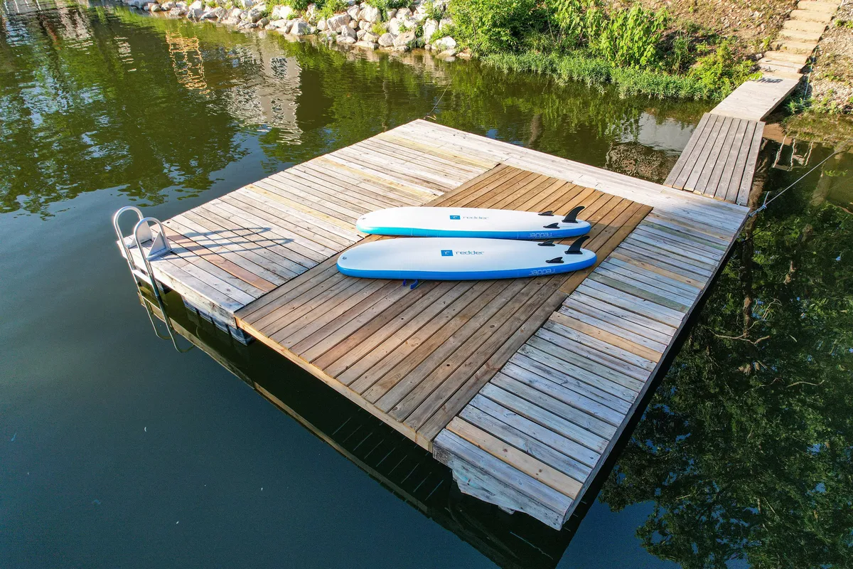 This image captures a wooden dock extending into the lake, with two brightly colored paddleboards ready for use. A ladder descends into the water, and lush greenery frames the scene, evoking an inviting and serene lakeside atmosphere.