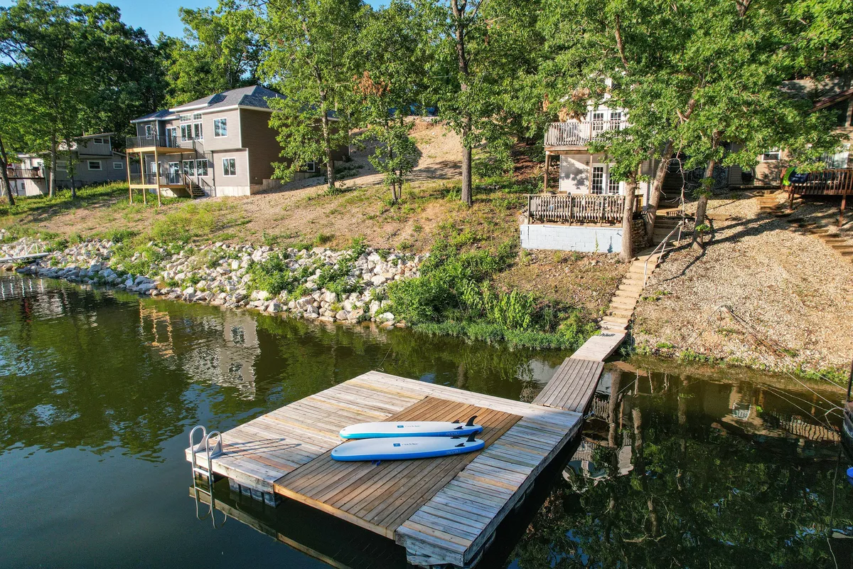 The image features a dock with kayaks ready for paddleboarding, surrounded by lush greenery and views of houses on the hillside. The tranquil water and accessible shoreline evoke possibilities for sunrise paddleboarding, relaxed water activities, and easy access to lake adventures.