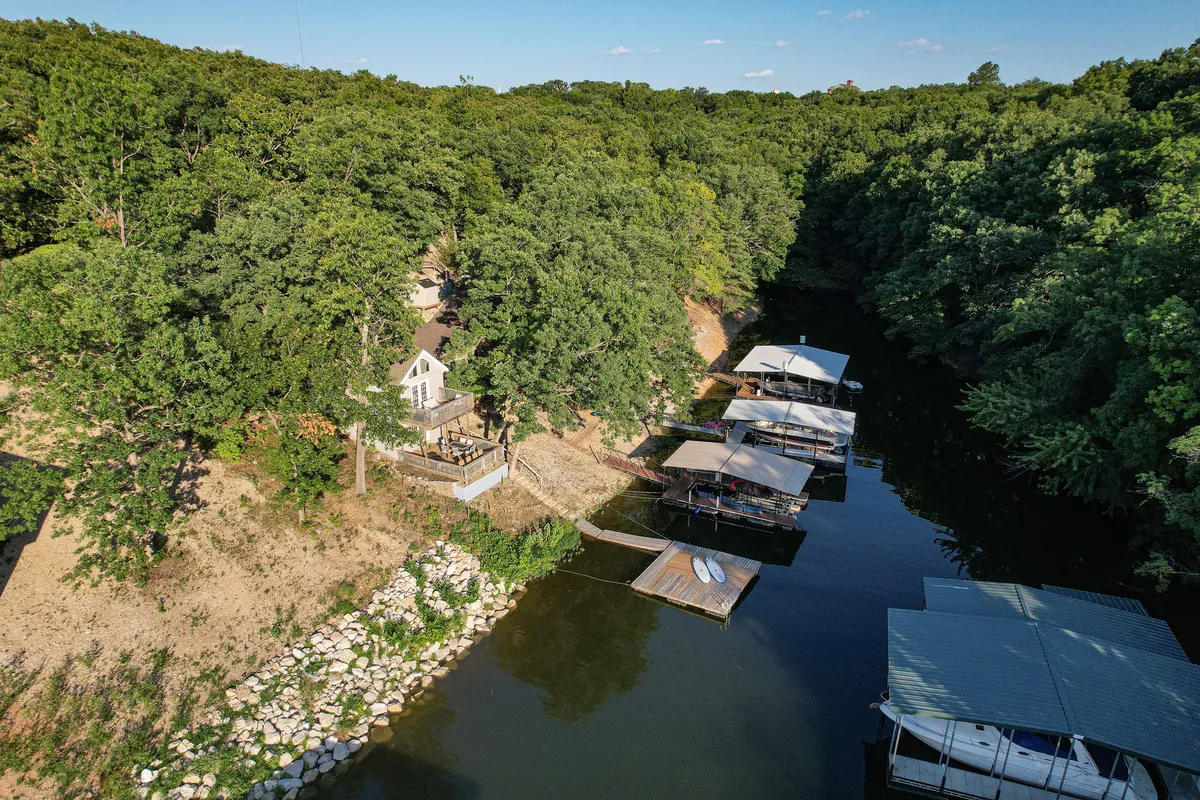 A wide-angle view displays the house perched above the dock, surrounded by trees, with boats on the water below. The setting radiates a sense of community and adventure, showing how close the accommodation is to the lake and inviting guests to explore both water and land.