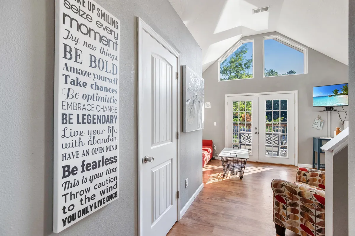 A stylish, sunlit sitting area with motivational wall art, a red seating area, and French doors leading outside. The space is bright and artistic, designed for relaxation, socializing, or enjoying morning coffee.