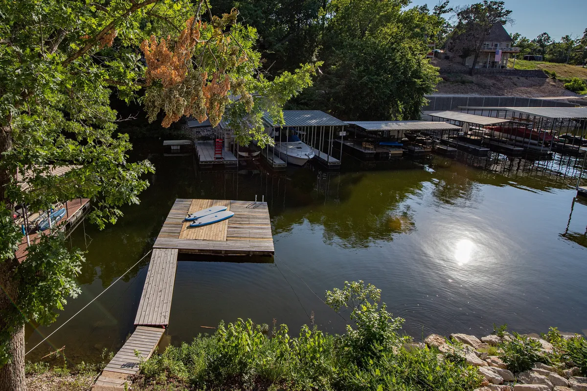A serene dock extends over the water, surrounded by lush trees with boats moored nearby. The space is inviting for outdoor relaxation, with ample room for enjoying the lakeside atmosphere and easy access for activities like fishing, boating, or letting a pet splash at the water’s edge.