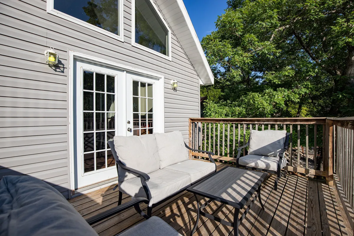 A welcoming exterior view of the lakefront home featuring an inviting wooden deck with outdoor seating. Large windows allow morning light to pour into the space, and the deck is elevated to give a panoramic view of the lake, perfect for enjoying coffee or intimate conversations. Lush greenery surrounds the home, reinforcing its private, romantic ambiance.