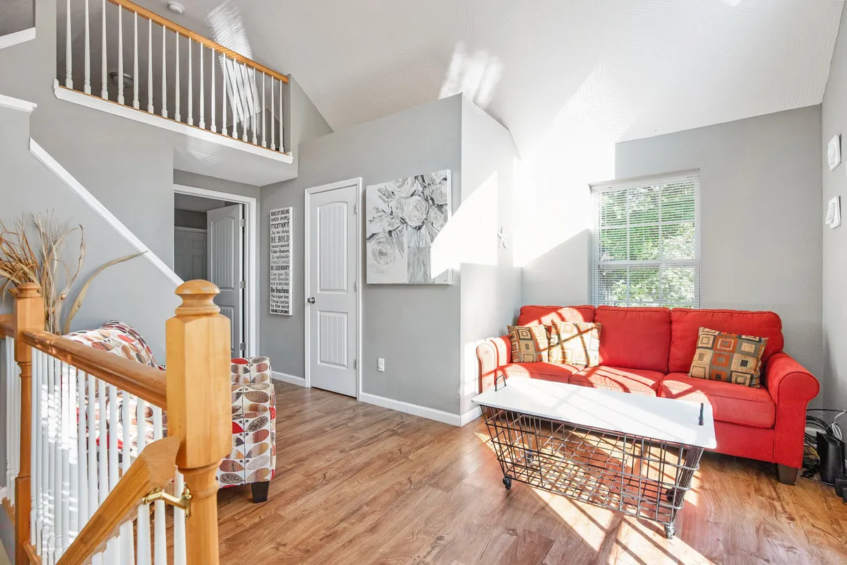 A welcoming living room featuring a red sofa, coffee table, and wall artwork, with a staircase visible in the background. Sunlight streams in, and the open layout makes the space inviting for gatherings or relaxing moments.