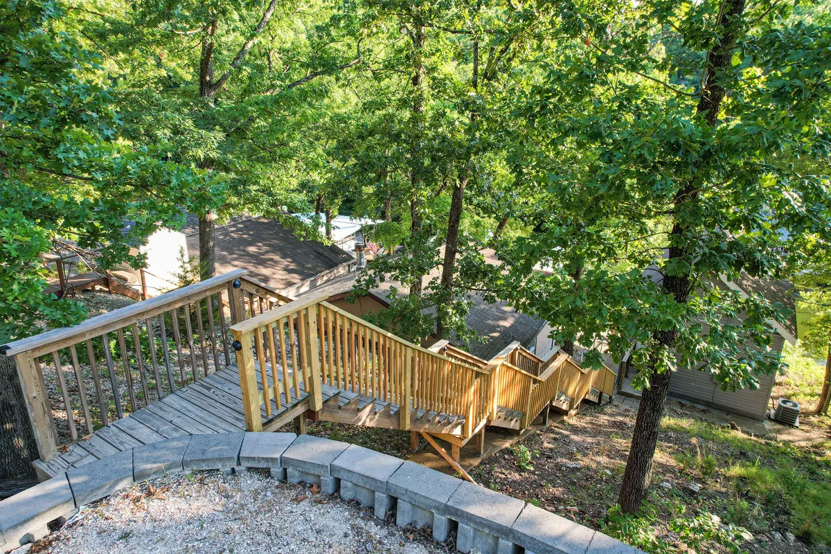 A rustic wooden staircase leads down through a grove of trees, with nearby cabins peeking through the greenery. The setting is peaceful and ideal for morning walks, nature observation, or gathering with family, including pets.