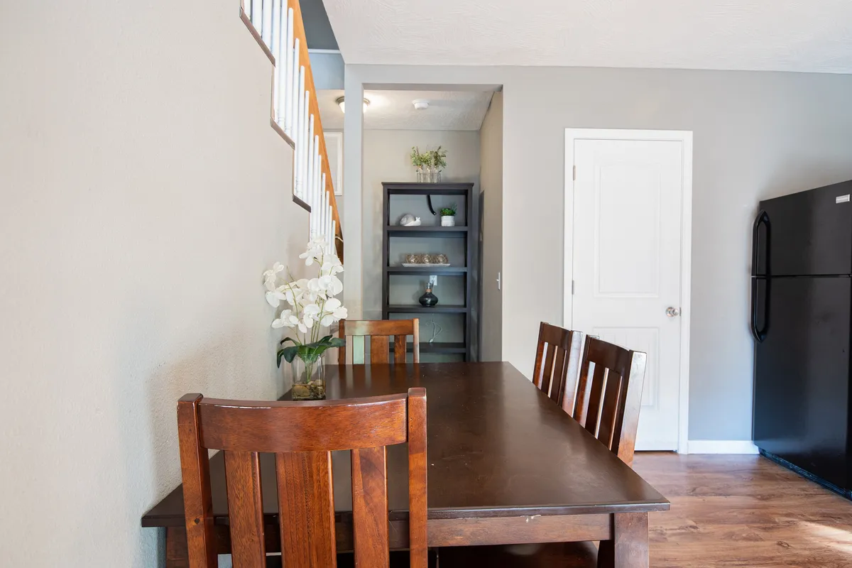 A cozy dining area is shown with a dining table, decorative shelving, a refrigerator, and an inviting staircase in the background. The space feels warm and perfect for family meals or social gatherings.