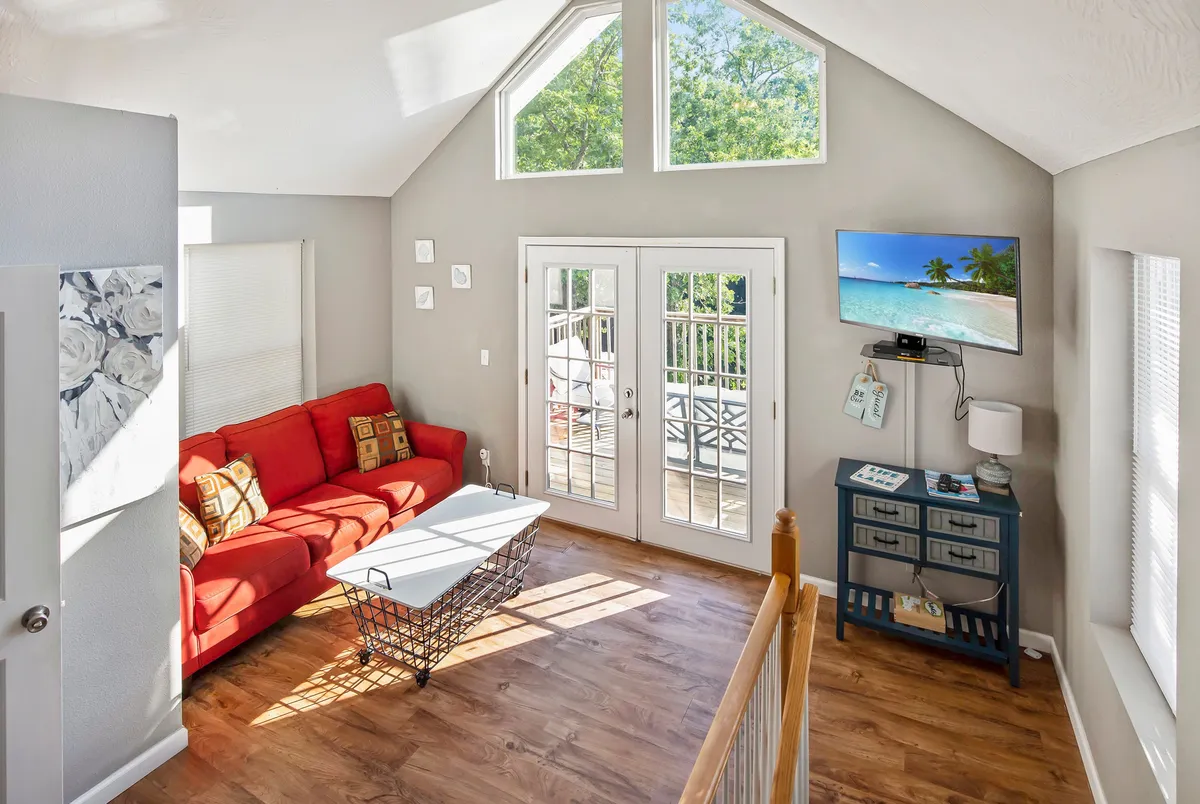 A cheerful living area with a plush red sofa facing a large TV, flooded with natural light streaming through glass doors. The space feels lively and inviting, perfect for relaxing with loved ones or pets. Hardwood floors and open sightlines contribute to a welcoming, homey atmosphere.